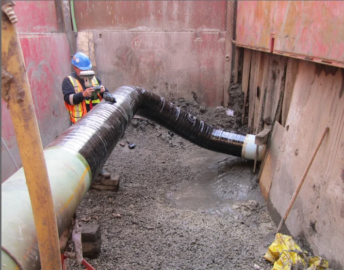 Worker inspecting a field-wrapped pipe coating in an excavation